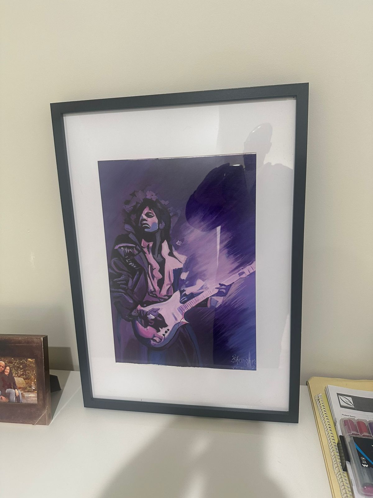A framed painting of a guitarist mid-solo, in violet and plum, standing on a white desk.